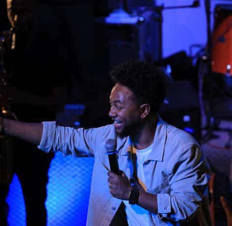 Zeruabbel Molla on stage holding a microphone and pointing towards the audience, with musical instruments in the background.