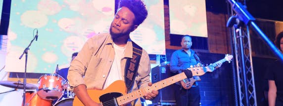 Zeruabbel Molla playing electric guitar on stage with colorful lights and screens in the background.