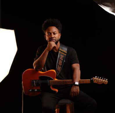 Zeruabbel Molla sitting on a stool holding an electric guitar, surrounded by studio lighting.