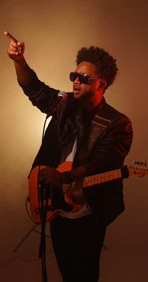 Zeruabbel Molla wearing sunglasses and a leather jacket, playing an electric guitar and pointing upwards.