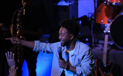 Zeruabbel Molla on stage with a microphone points towards the audience on stage, with musical instruments in the background.