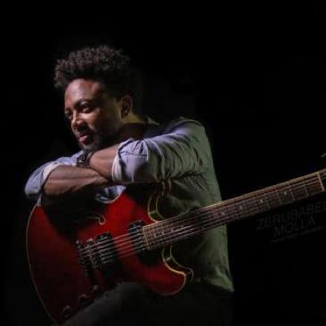 Zerubabbel Molla sitting with a red electric guitar in a dark setting.